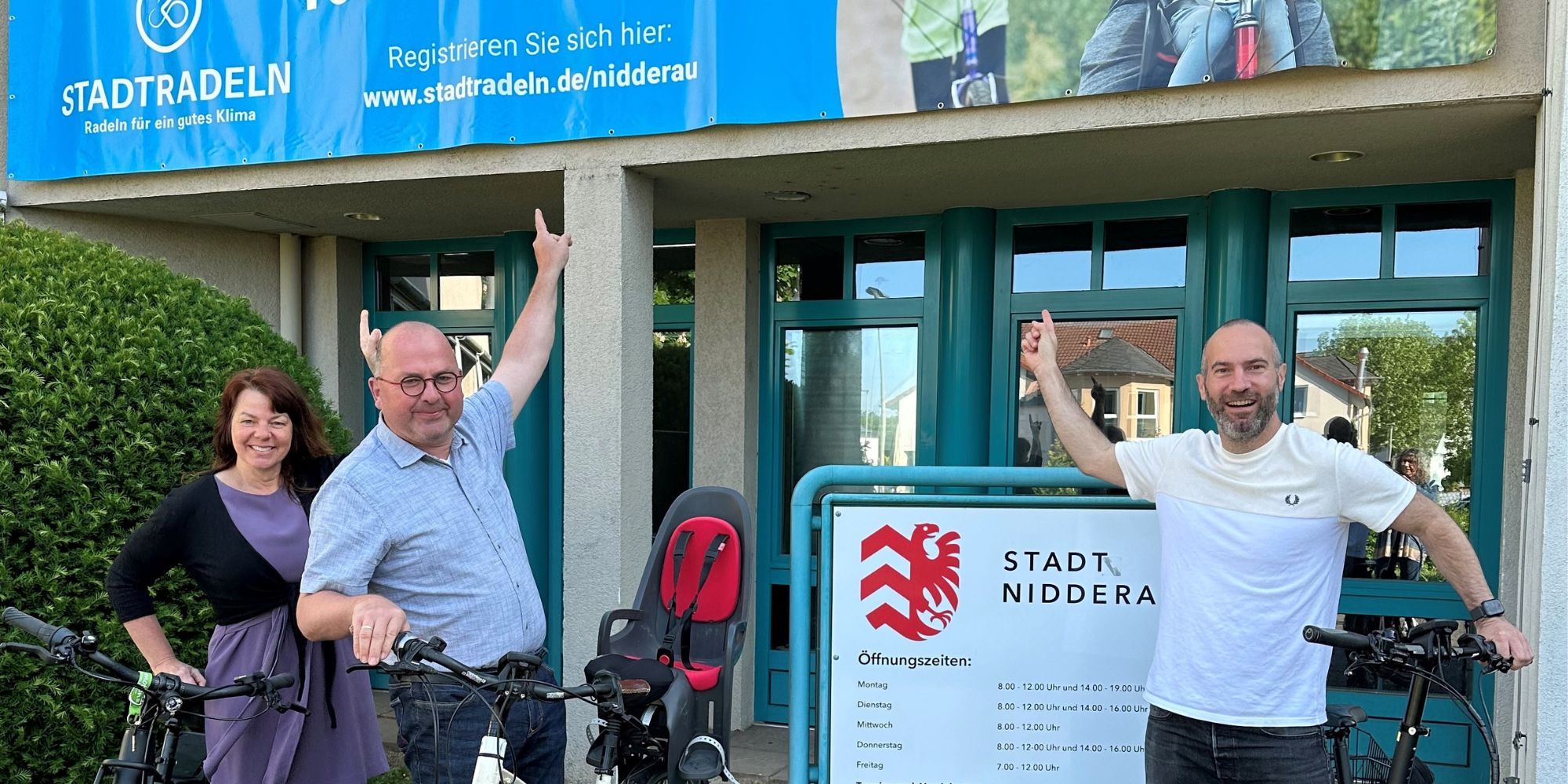 Katja Adams, Rainer Vogel und Andreas Bär zeigen auf das Stadtradeln-Banner am Rathaus