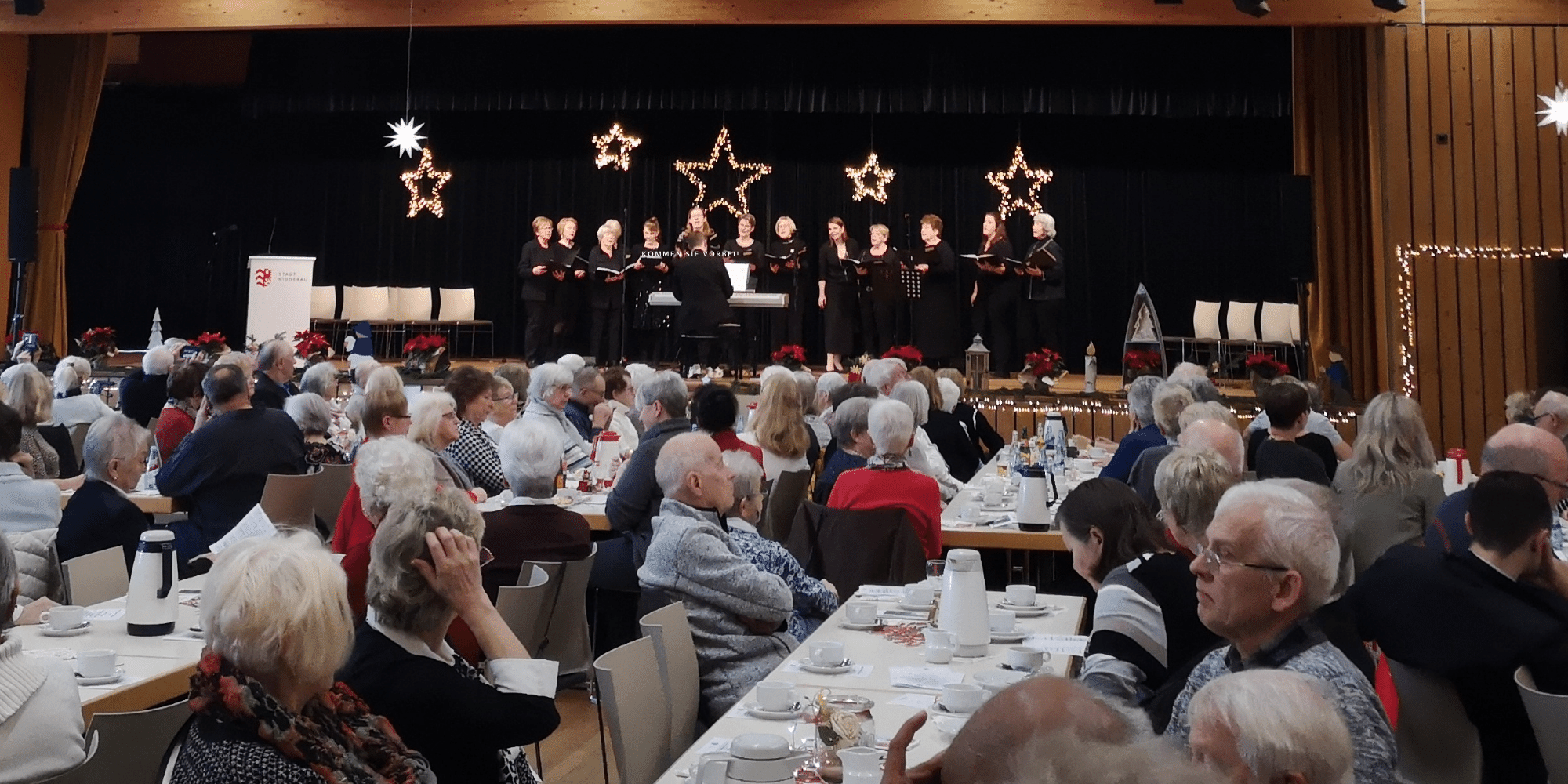 Mehrere ältere Personen sitzend an Tischen vor einer schwarzen Bühne