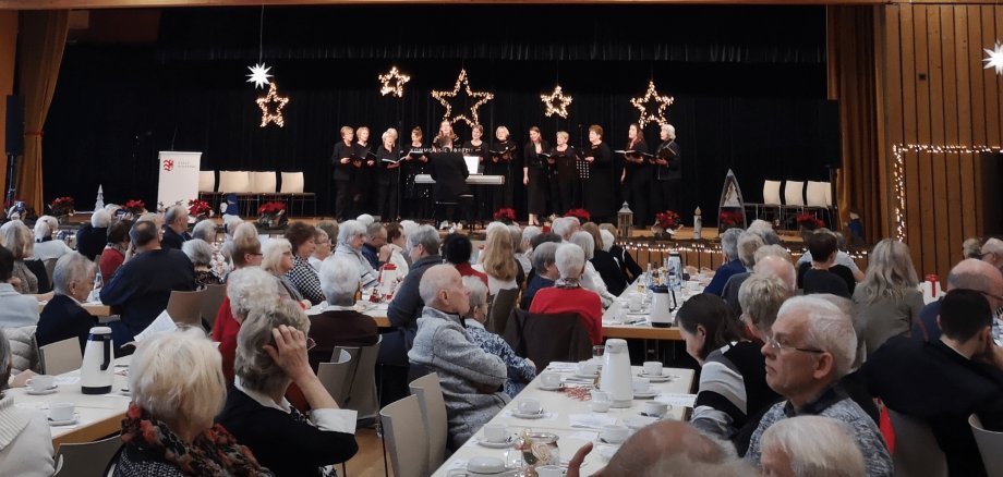 Mehrere ältere Personen sitzend an Tischen vor einer schwarzen Bühne