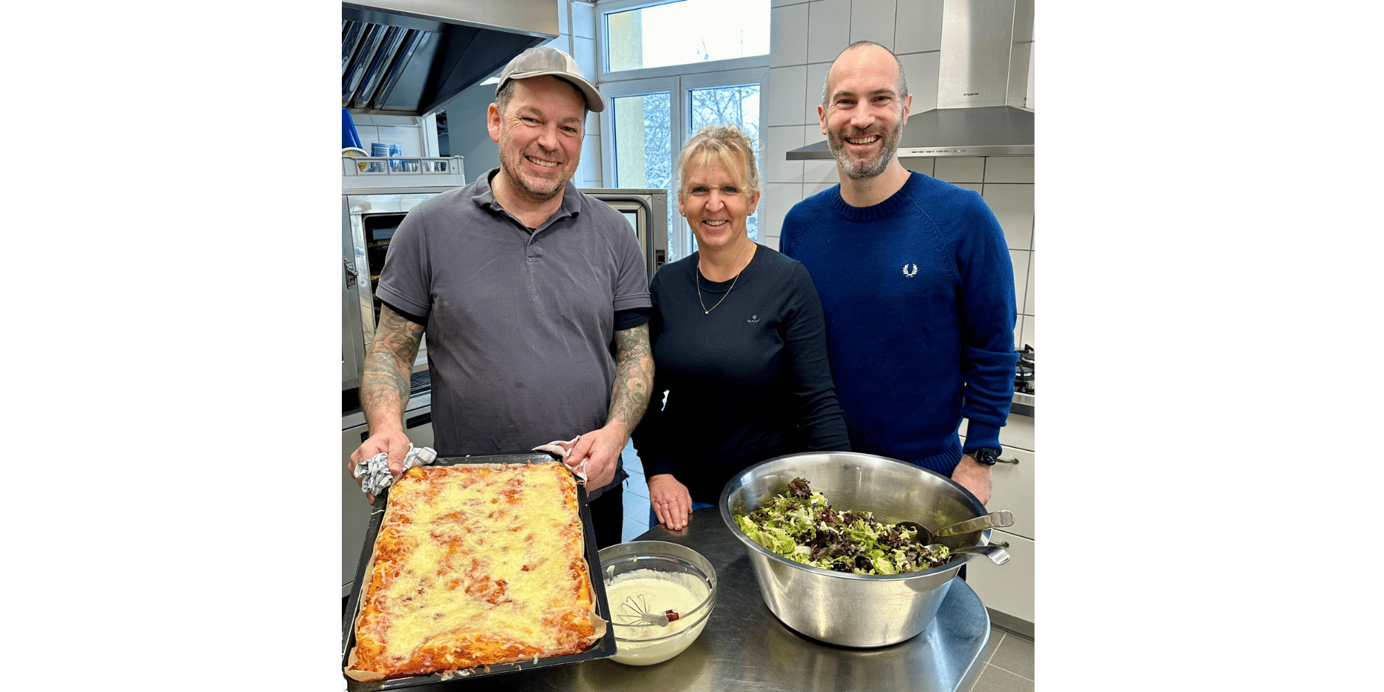 Drei Personen stehend in einer  Küche vor einem Tisch mit Salat und Pizza Margherita