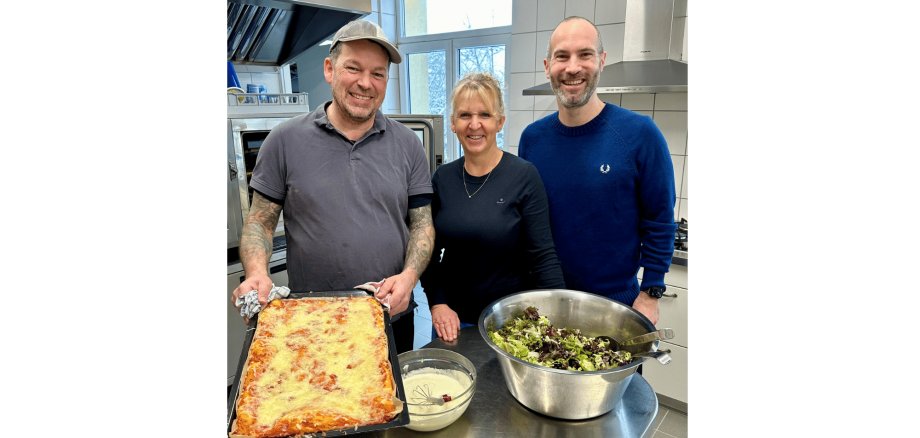 Drei Personen stehend in einer  Küche vor einem Tisch mit Salat und Pizza Margherita
