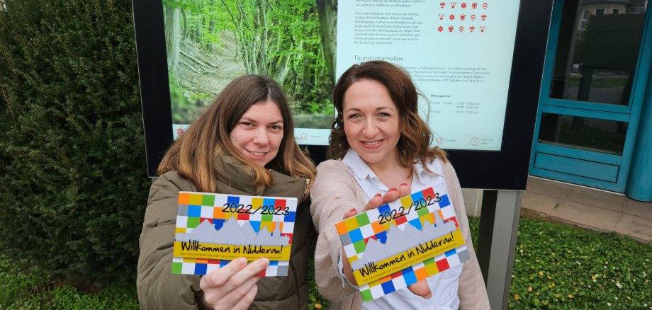 Zwei Frauen stehend vor dem digitalen Display des Rathauses und in der Hand haltend jeweils das Gutscheinheft der Stadt Nidderau
