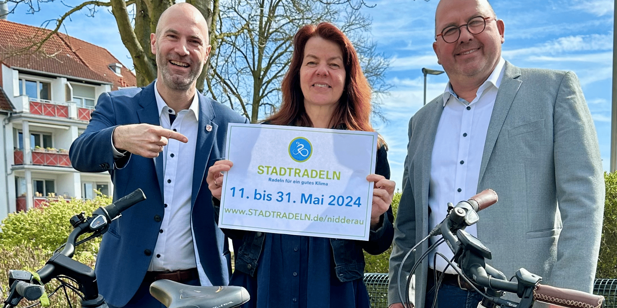 2 Männer und 1 Frau stehend hinter 2 Fahrrädern. Die Frau hält ein Plakat mit der Aufschrift "Stadtradeln" hoch