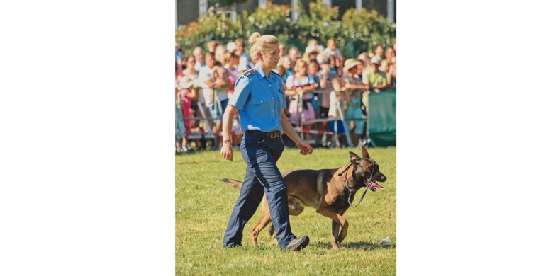 Frau in Polizeiuniform mit einem Schäferhund läuft über einen Rasen umgeben von Zuschauern