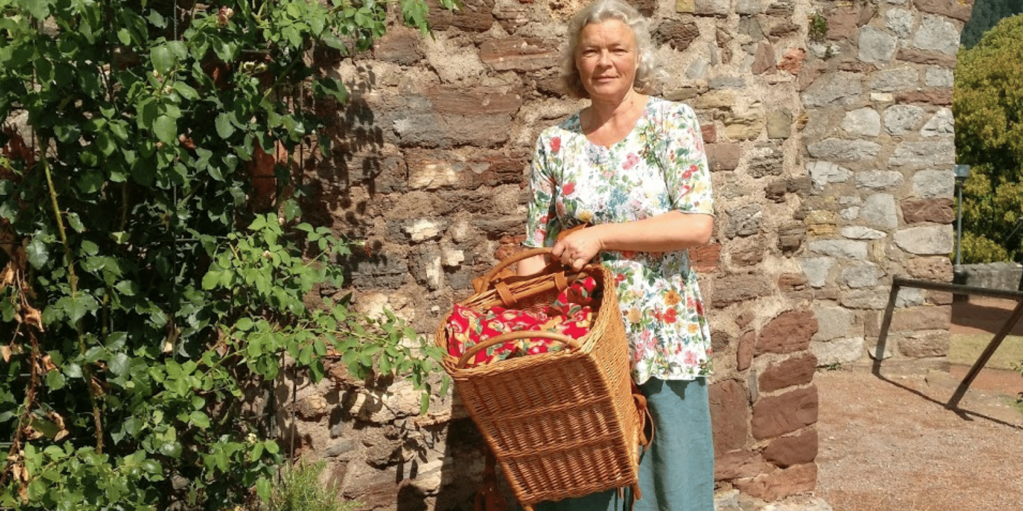 Frau mit weißen Haaren und geblümtem Shirt und Flechtkorb steht vor einer Steinmauer, welche links mit Efeu bewachsen ist
