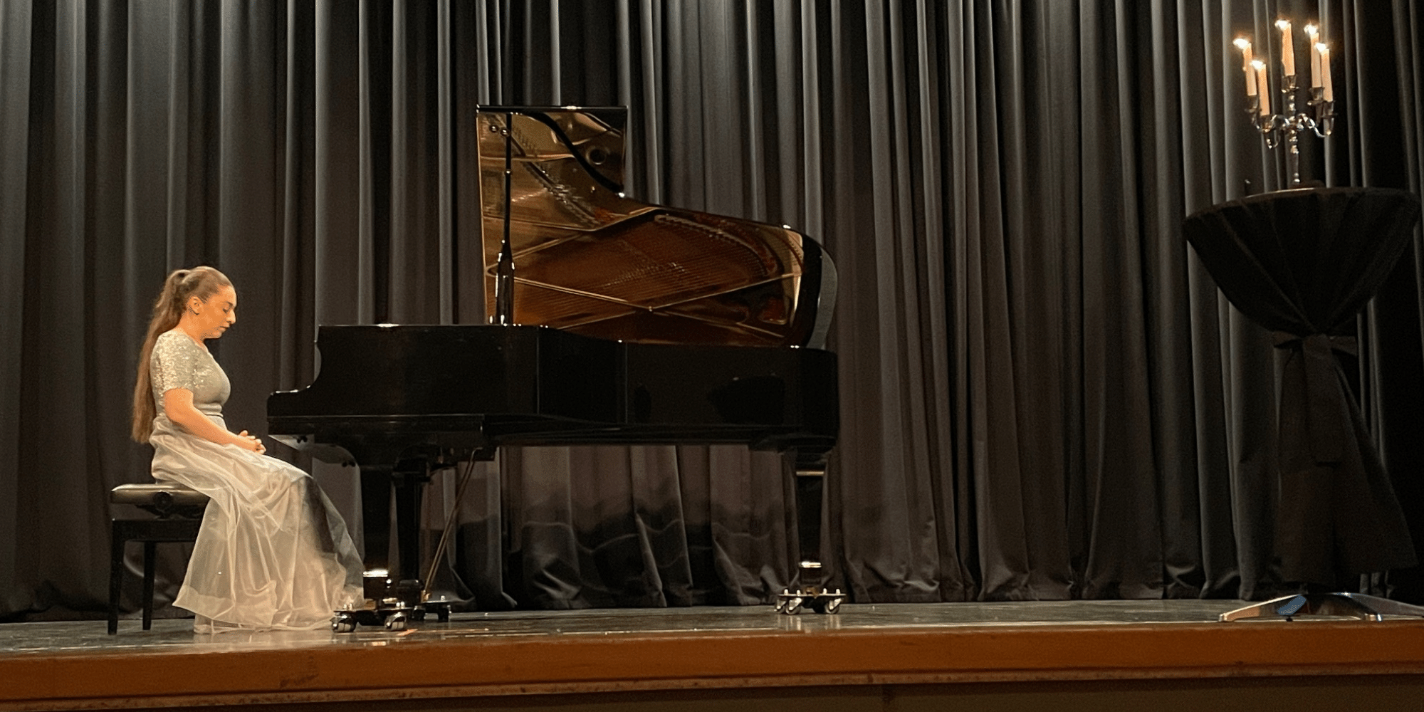 Frau mit langen braunen Haaren und einem Kleid, sitzend am Klavier auf einer schwarzen Bühne, auf einem Tisch weiter rechts steht ein angezündeter Kerzenständer