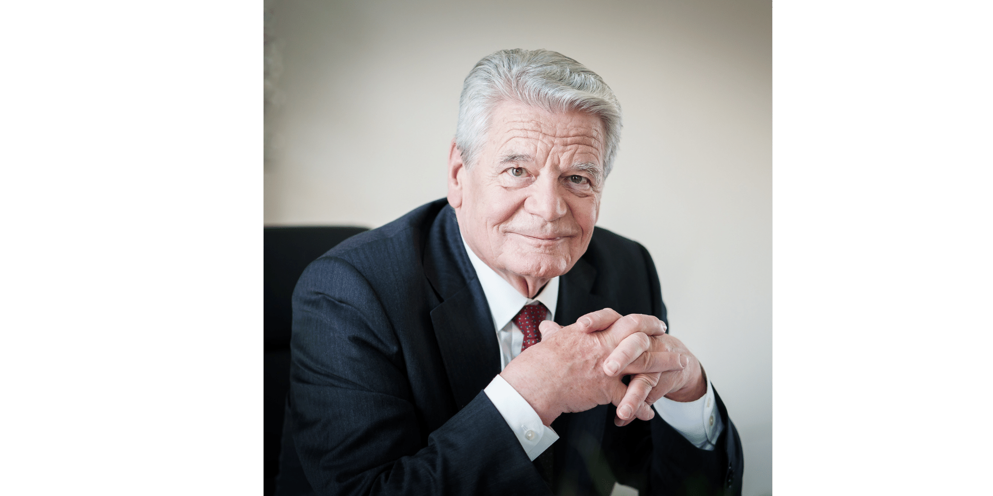 Mann mit weißen Haaren im dunklen Anzug sitzend mit den Ellenbogen aufstützend auf einem Tisch und die Hände gefaltet