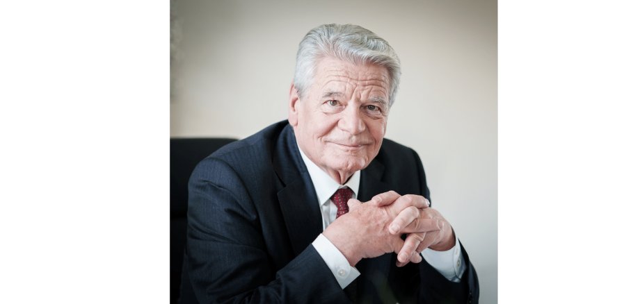 Mann mit weißen Haaren im dunklen Anzug sitzend mit den Ellenbogen aufstützend auf einem Tisch und die Hände gefaltet