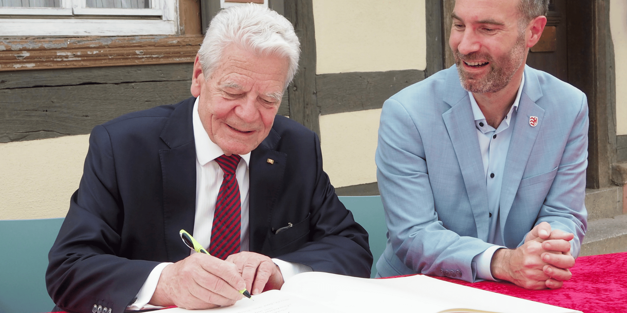 Zwei Männer in Anzügen sitzend vor einem Tisch mit einem großen Buch davor. Im Hintergrund ist ein Fachwerkhaus
