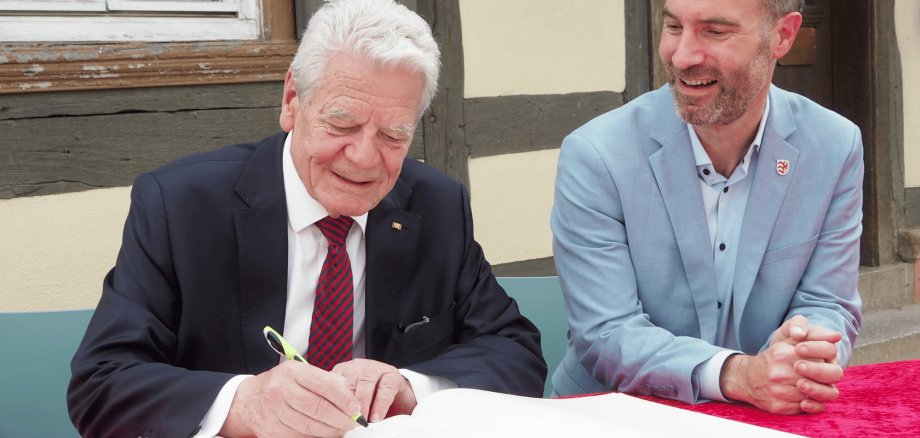 Zwei Männer in Anzügen sitzend vor einem Tisch mit einem großen Buch davor. Im Hintergrund ist ein Fachwerkhaus
