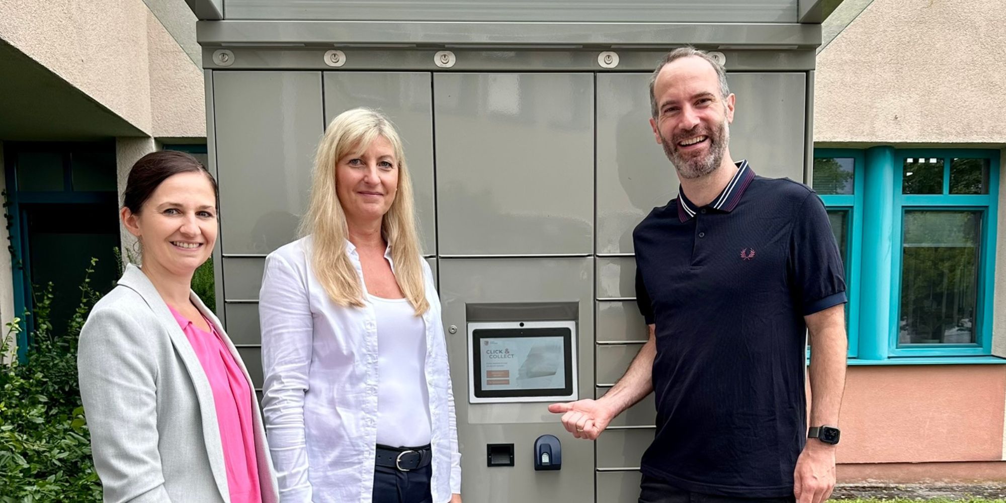 Zwei Frauen und ein Mann zeigen auf einen grauen Automaten