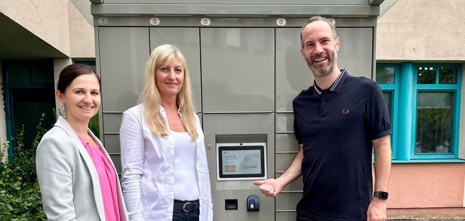 Zwei Frauen und ein Mann zeigen auf einen grauen Automaten