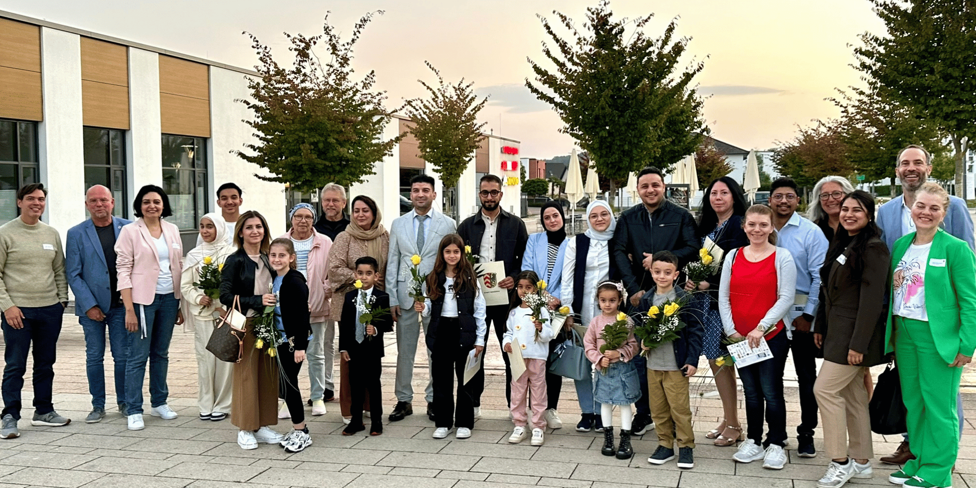 Mehrere Personen stehend auf einem Platz. Manche Personen halten gelbe Rosen in der Hand