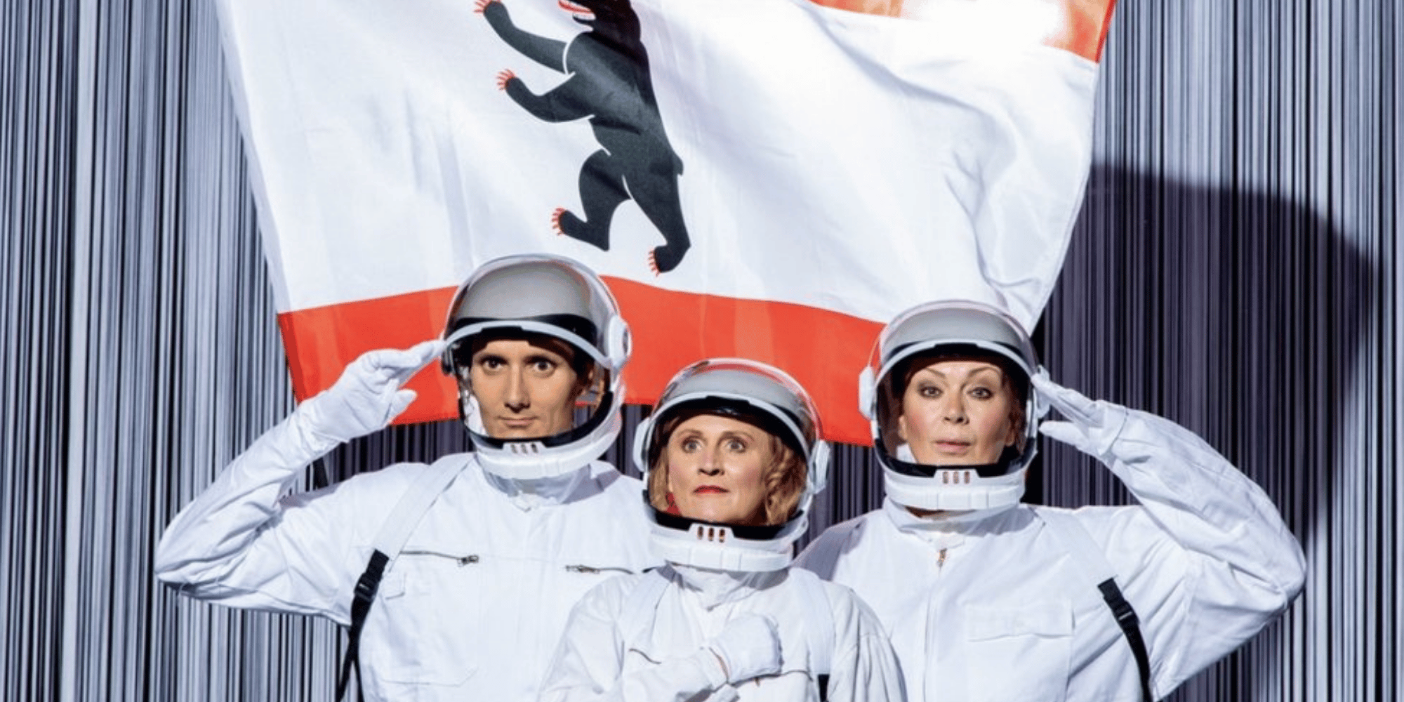 2 Frauen und 1 Mann in weißen Astronautenanzügen vor einer Berliner Flagge