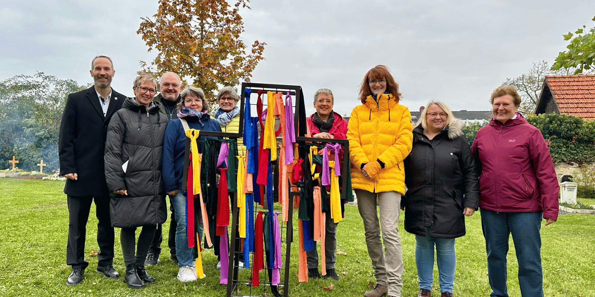 Mehrere Personen stehend auf einem Friedhof mit mehreren bunten Bändern