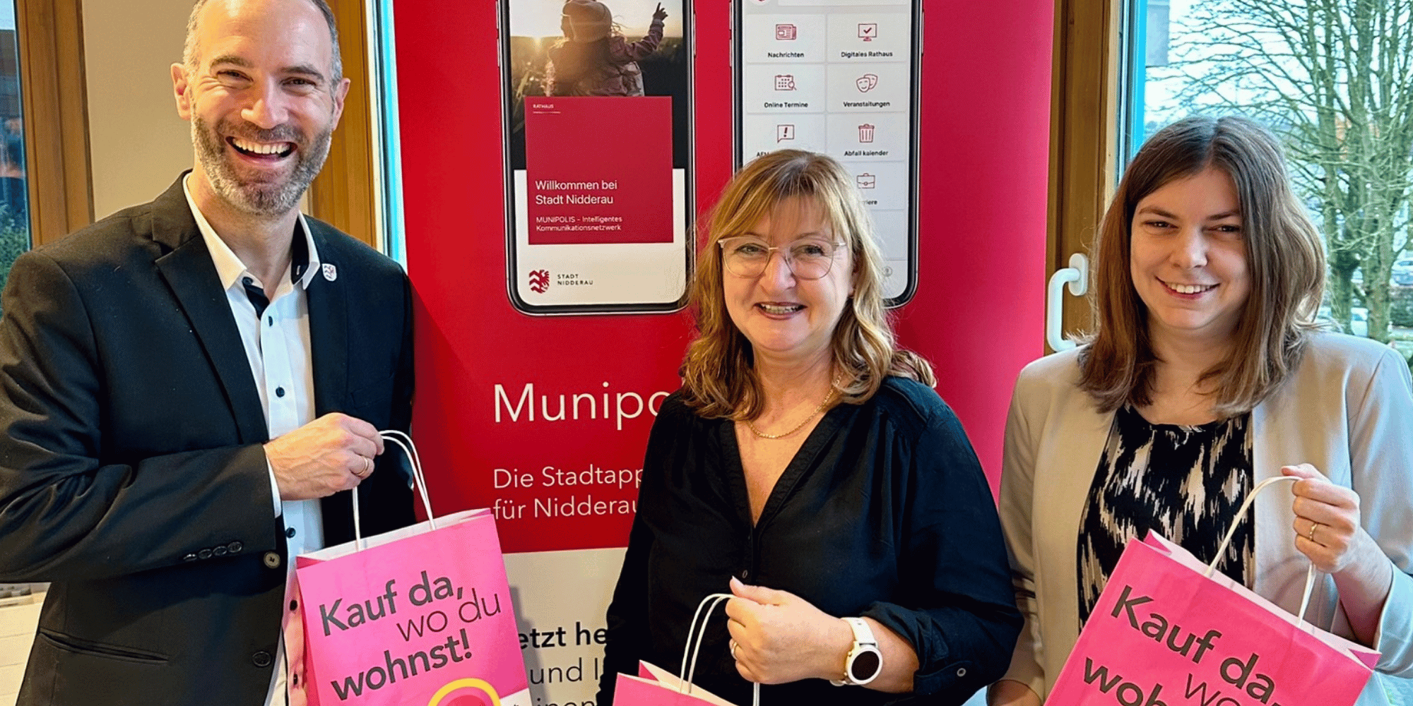 Zwei Frauen und ein Mann halten rosa-farbene Taschen und stehen vor einem roten Banner