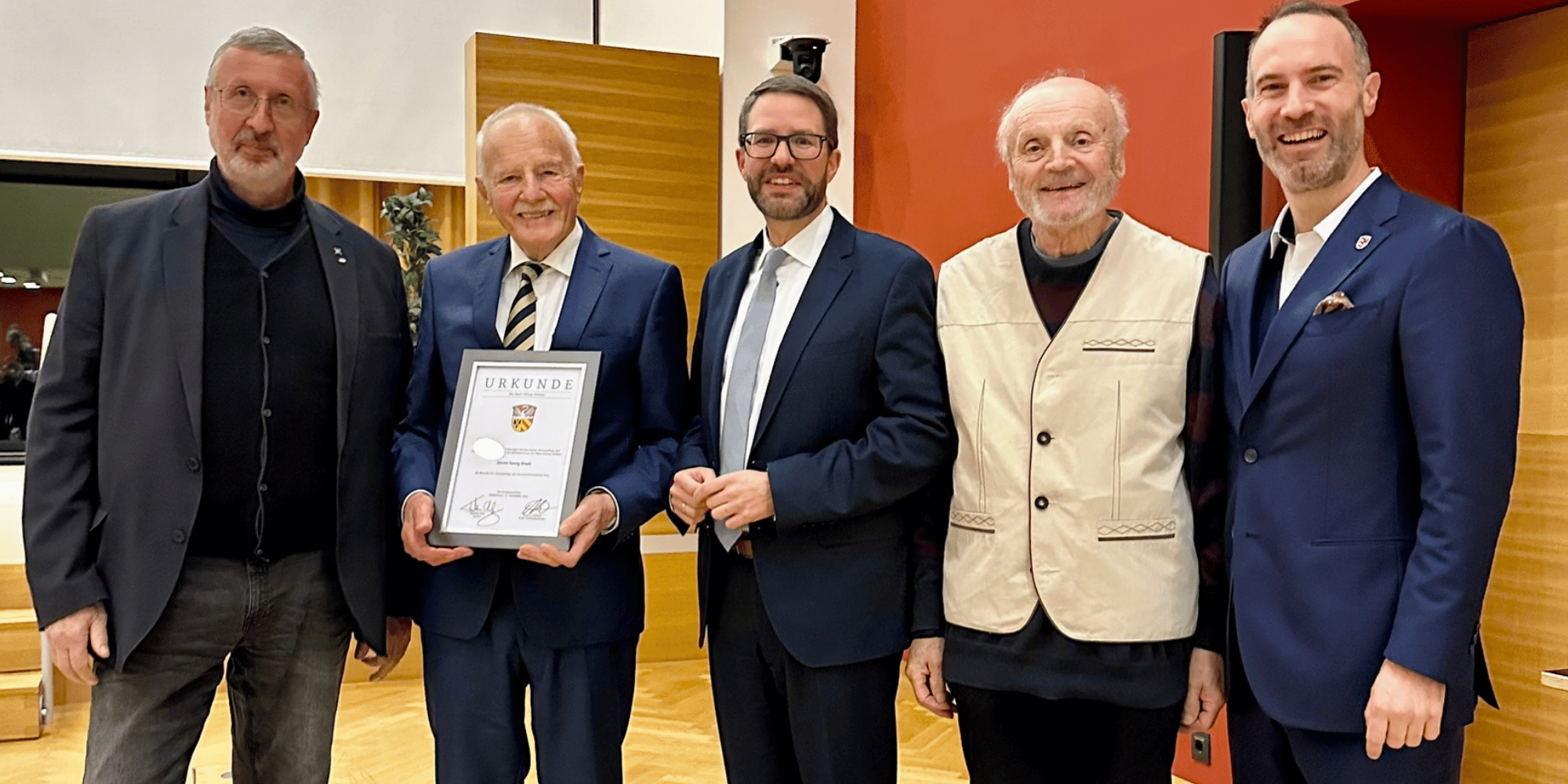 Fünf Männer in Anzügen stehend. Ein Mann hält eine Urkunde vor sich