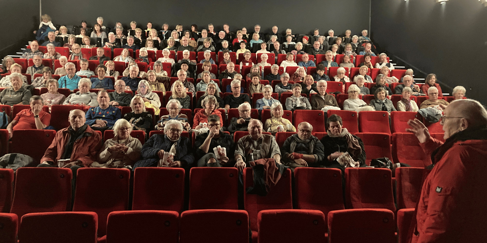 Kinosaal mit vielen besetzten Plätzen. Im Vordergrund rechts steht ein Mann in roter Jacke