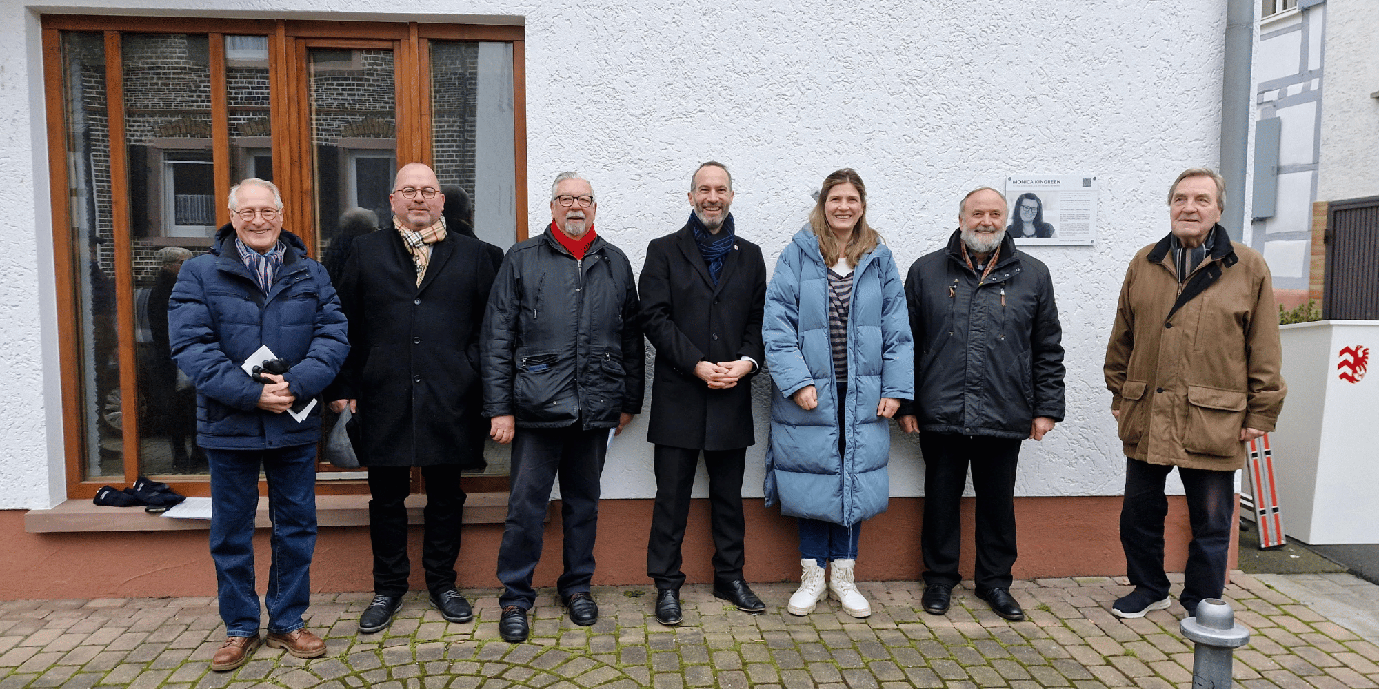Sechs Männer und eine Frau stehend vor der Hausfassade eines weißen Hauses mit dunklem Schriftzug "Monica Kingreen Haus"