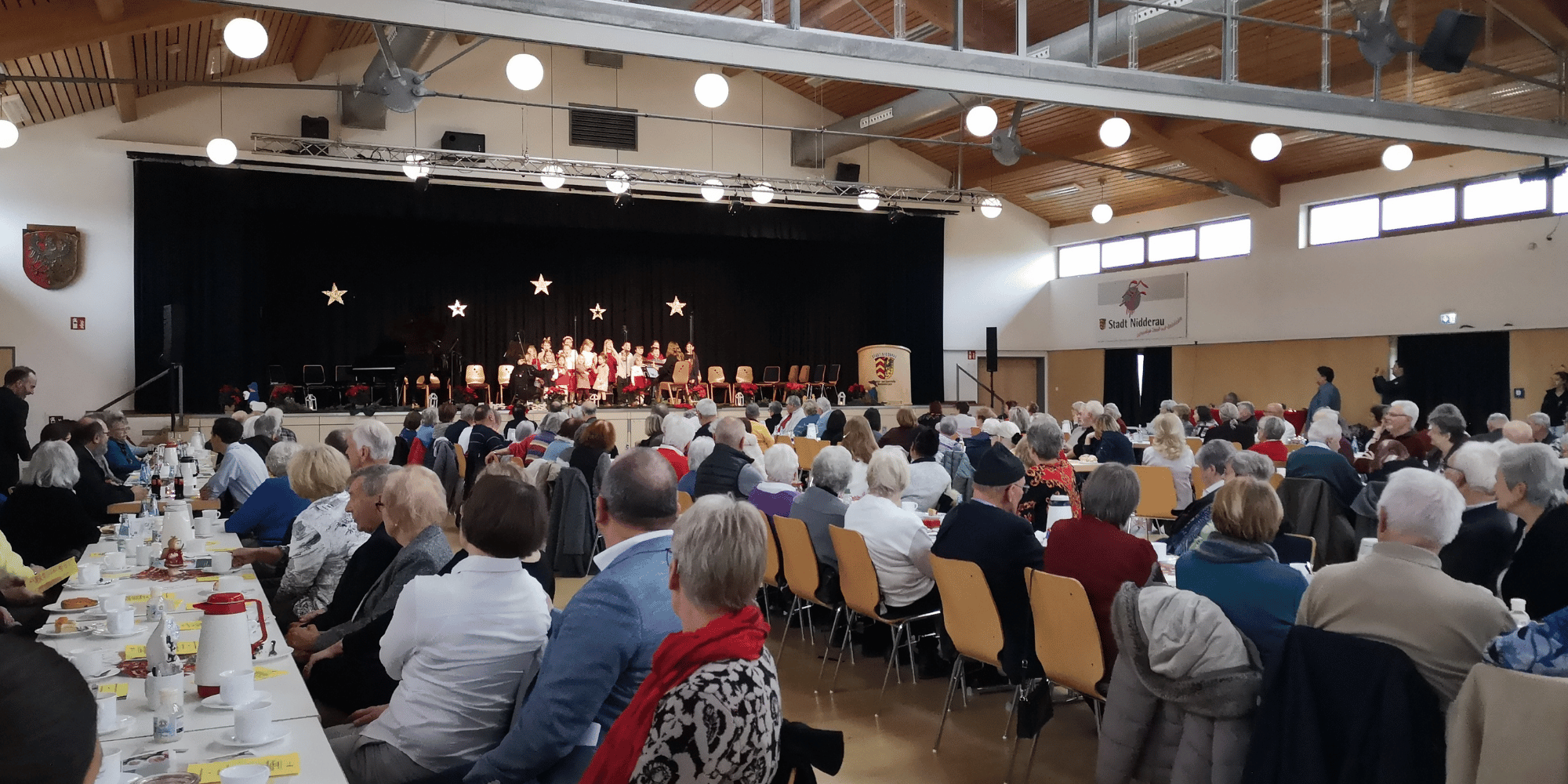 Mehrere Personen sitzend an Tischen und auf eine Bühne schauend