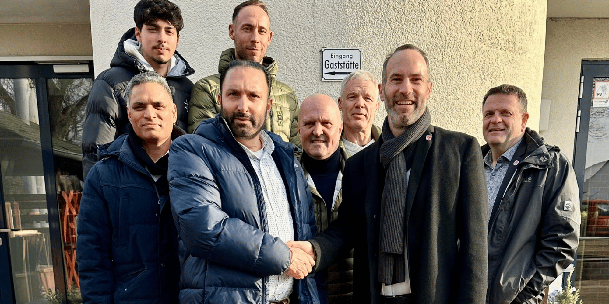 Acht Männer in Jacken stehend vor einem Haus. Zwei Männer geben sich die Hand