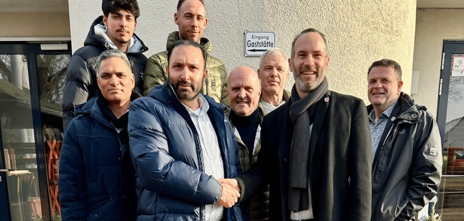 Acht Männer in Jacken stehend vor einem Haus. Zwei Männer geben sich die Hand