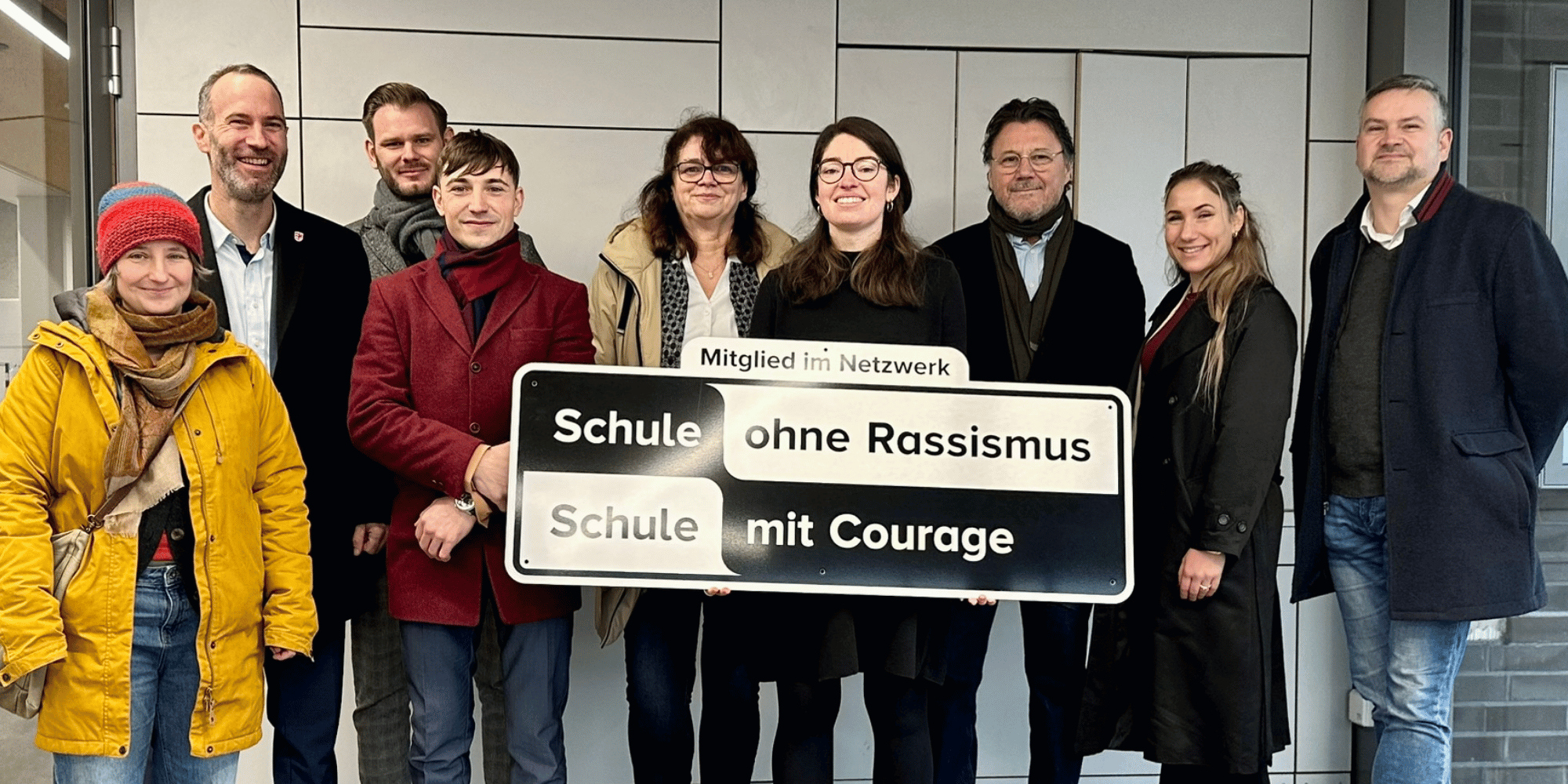 Mehrere Personen stehend. Vorne wird ein schwarz-weißes Schild gehalten mit der Aufschrift "Mitglied im Netzwerk Schule ohne Rassismus, Schule mit Courage"