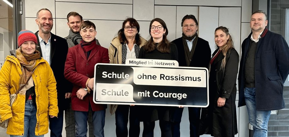 Mehrere Personen stehend. Vorne wird ein schwarz-weißes Schild gehalten mit der Aufschrift "Mitglied im Netzwerk Schule ohne Rassismus, Schule mit Courage"