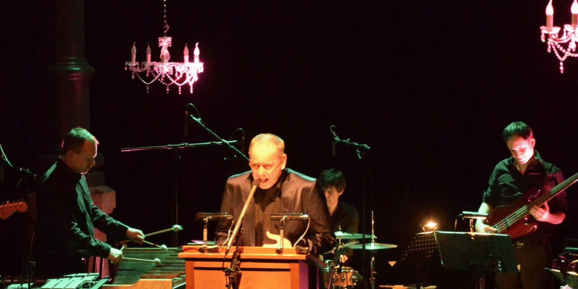 Mann in schwarzer Kleidung auf einer Bühne, an der Decke hängen Kronleuchter