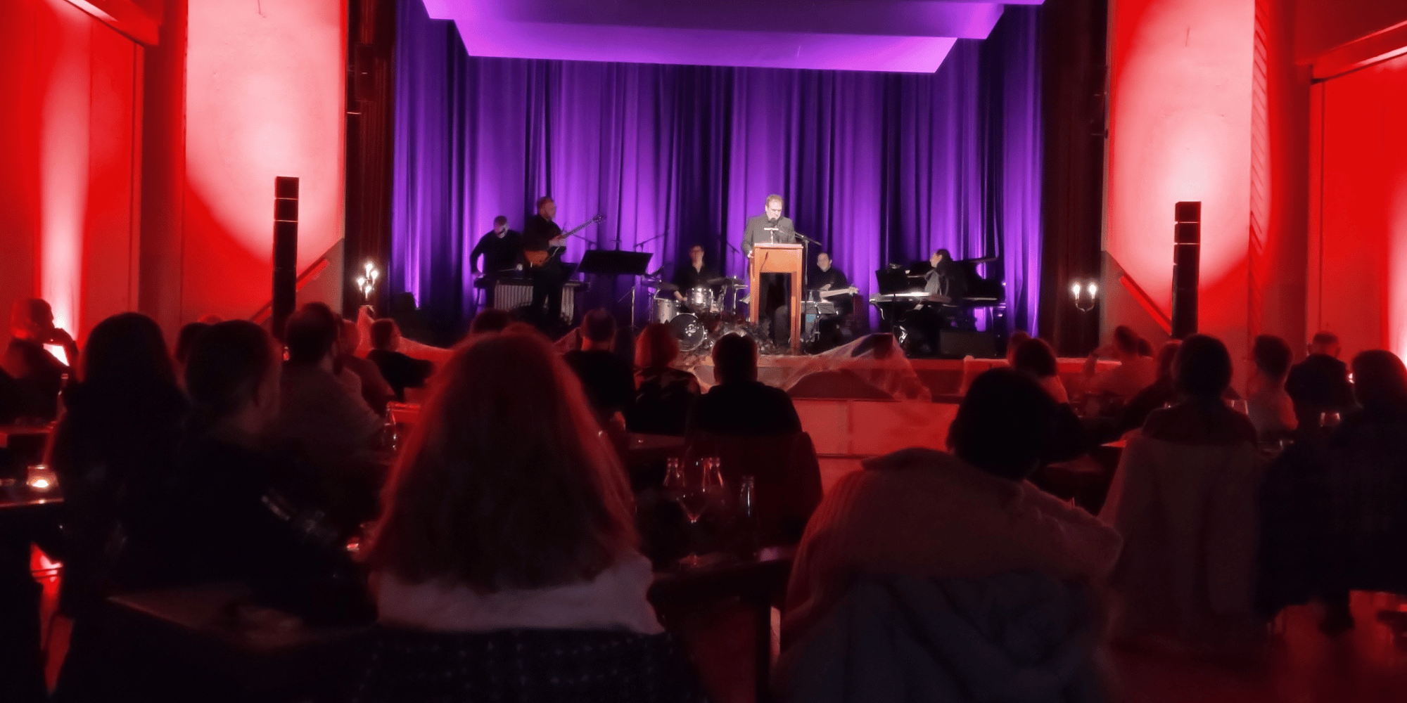 Mann in schwarzem Anzug auf einer rot-lila beleuchteten Bühne mit sitzendem Publikum davor