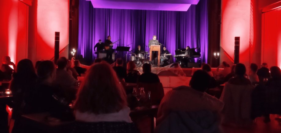 Mann in schwarzem Anzug auf einer rot-lila beleuchteten Bühne mit sitzendem Publikum davor