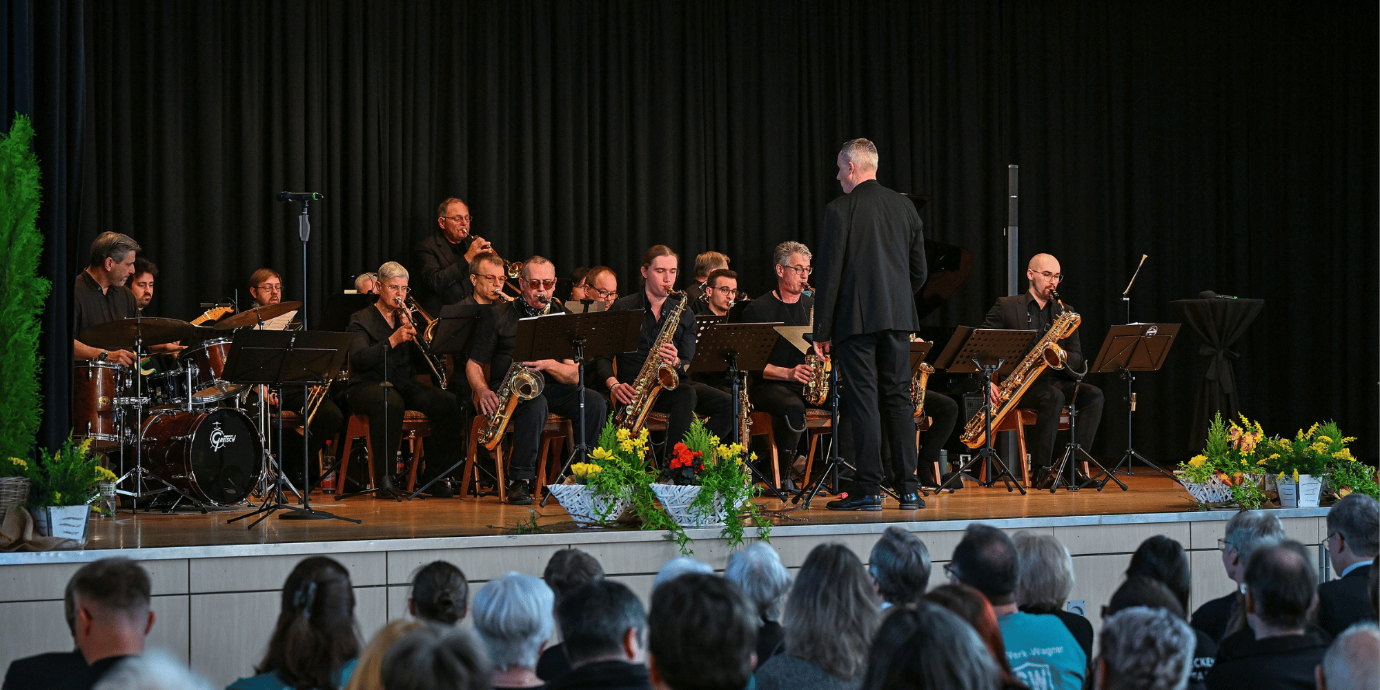Bühne sitzendem Orchester darauf. Die Musiker sind schwarz gekleidet 
