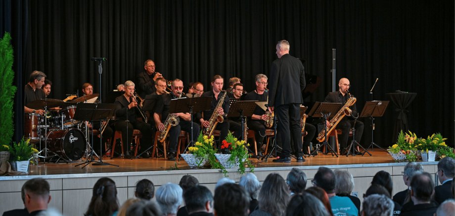 Bühne sitzendem Orchester darauf. Die Musiker sind schwarz gekleidet 