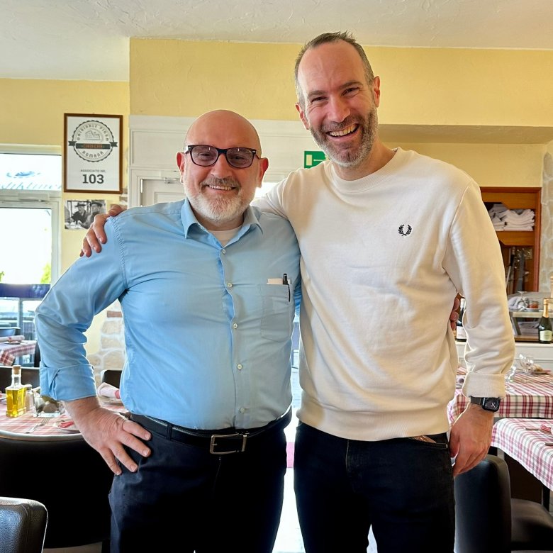 Zwei Männer stehend, der eine im blauen Hemd, der andere im weißen Pullover
