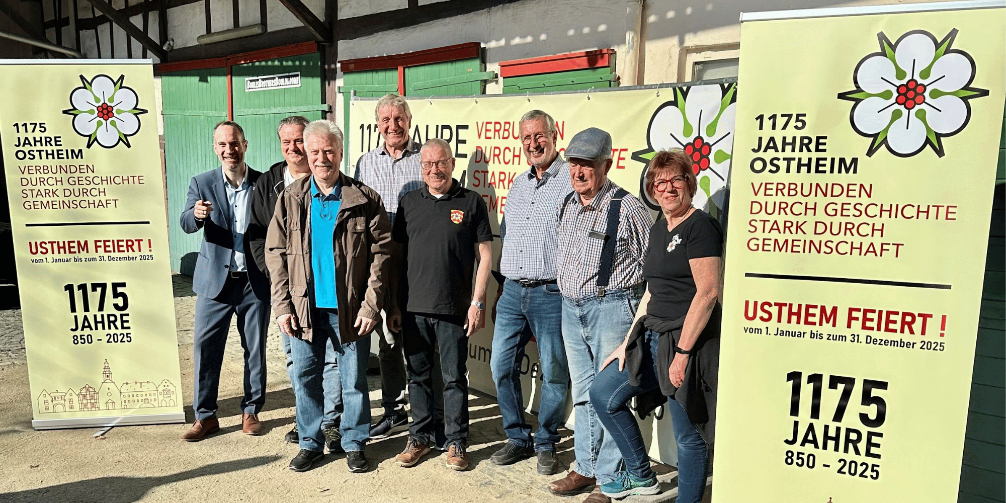 7 Männer und 1 Frau stehend in einem Fachwerkhof neben Roll-Ups mit dem Titel "1175 Jahre Ostheim"