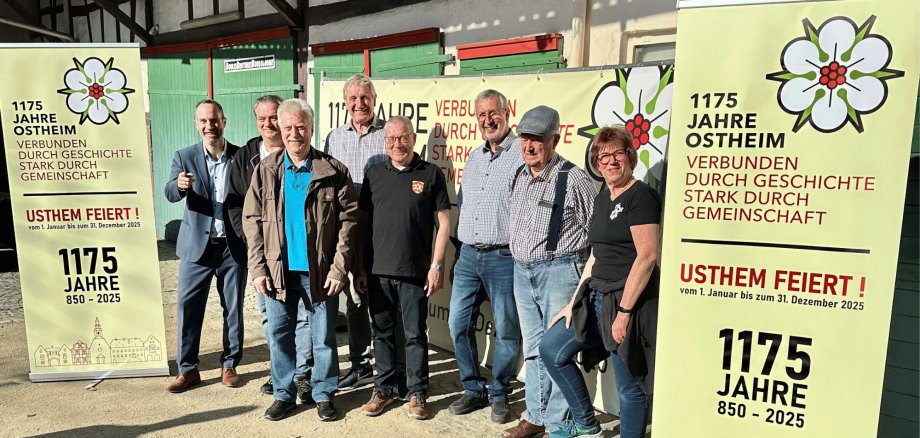 7 Männer und 1 Frau stehend in einem Fachwerkhof neben Roll-Ups mit dem Titel "1175 Jahre Ostheim"
