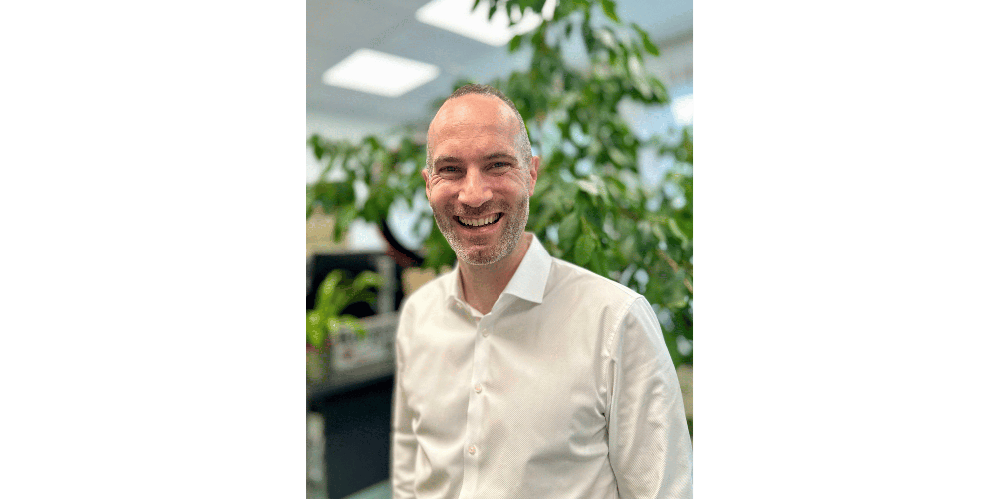Ein Mann im weißen Hemd lächelnd und stehend vor einer grünen Pflanze in einem Büro