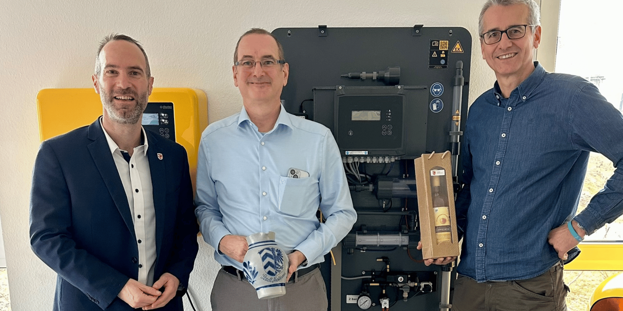 Drei Männer stehend vor einem elektronischen Schaltschrank. Ein Mann hält einen Bembel und ein anderer hält eine eingepackte Flasche in der Hand
