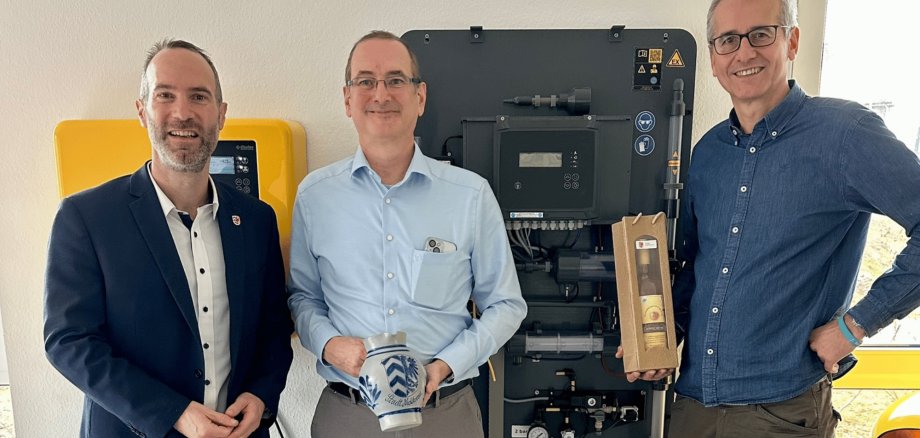 Drei Männer stehend vor einem elektronischen Schaltschrank. Ein Mann hält einen Bembel und ein anderer hält eine eingepackte Flasche in der Hand