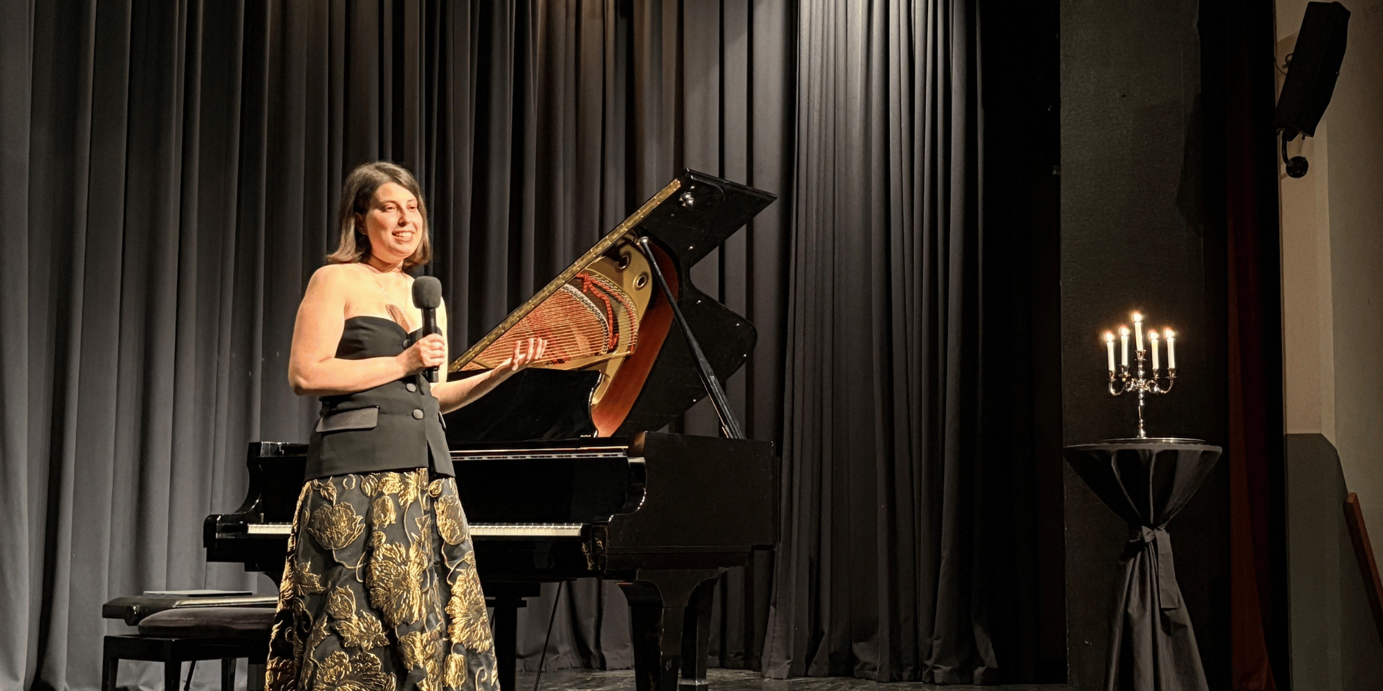 Eine Frau stehend auf einer schwarzen Bühne mit einem Fächer in den Hand haltend vor einem Klavier