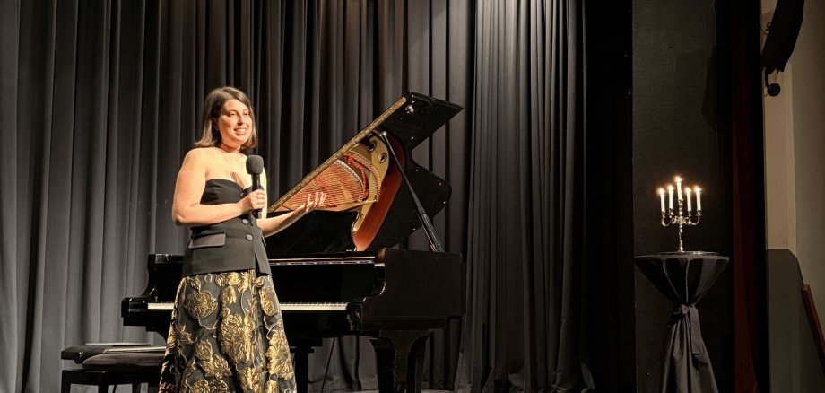 Eine Frau stehend auf einer schwarzen Bühne mit einem Fächer in den Hand haltend vor einem Klavier
