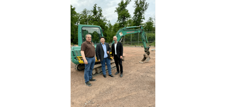 3 Männer stehend auf einer Baustelle eines Sportplatzes vor einem Bagger