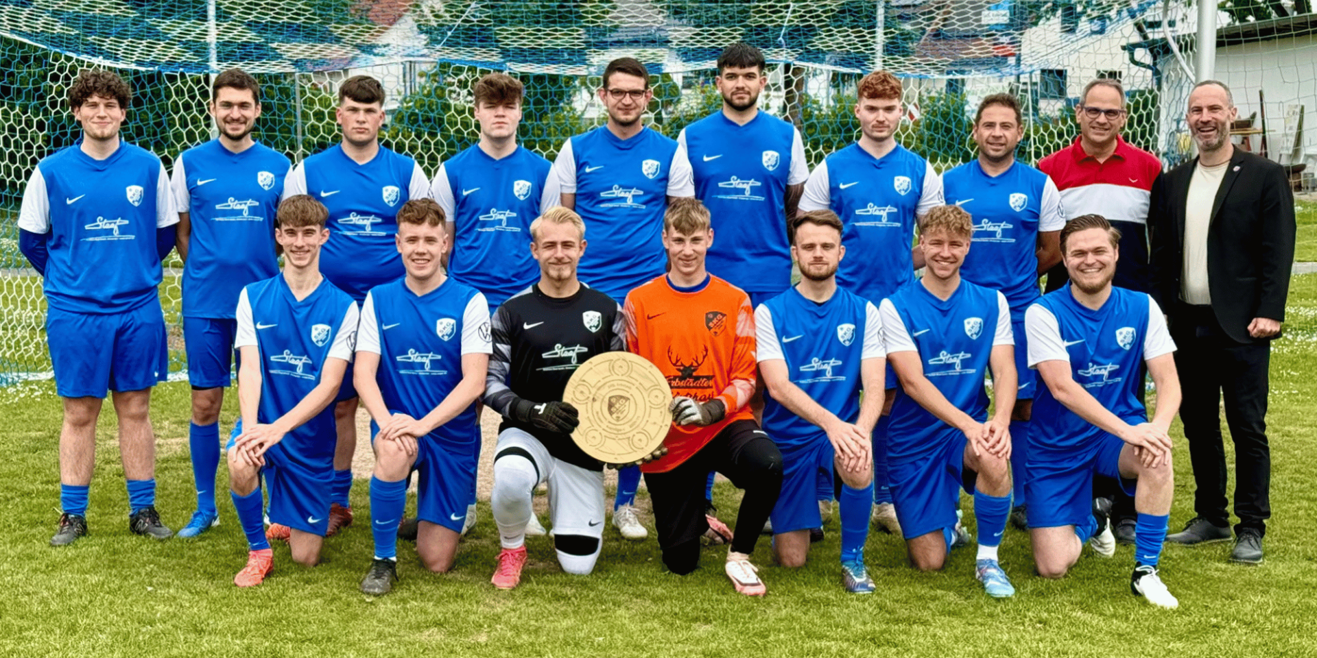 Fußballmannschaft bestehend aus mehreren Männern in blauen Trikots stehend vor einem Tor. In der Mitte halten zwei davon eine goldene Schale