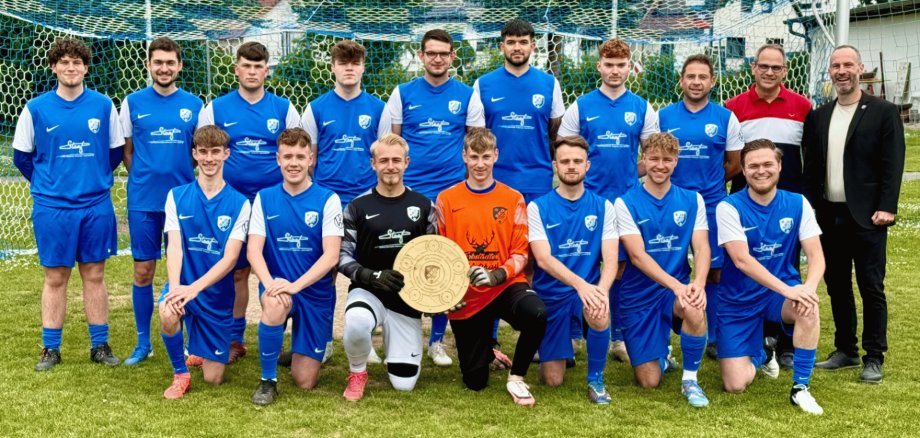 Fußballmannschaft bestehend aus mehreren Männern in blauen Trikots stehend vor einem Tor. In der Mitte halten zwei davon eine goldene Schale