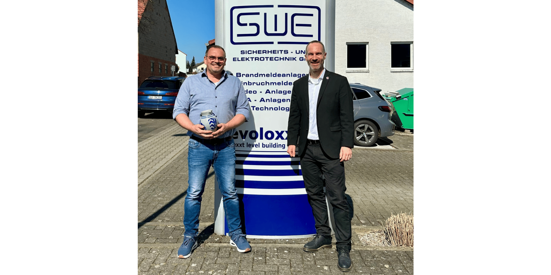 Zwei Männer stehend vor einem Firmenschild. der eine Mann hält einen Bembel  (hessischen Krug) in der Hand