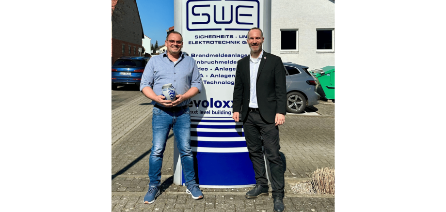 Zwei Männer stehend vor einem Firmenschild. der eine Mann hält einen Bembel  (hessischen Krug) in der Hand