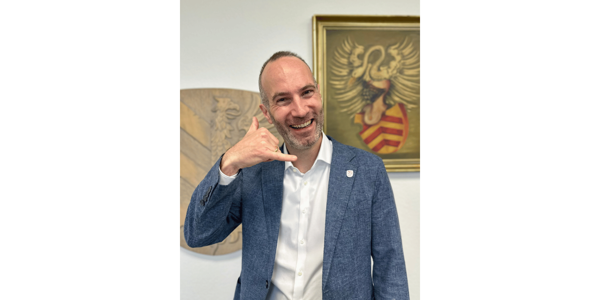 Mann in einem blauen Sakko lächelnd und stehend vor einem Gemälde mit einem Wappen.