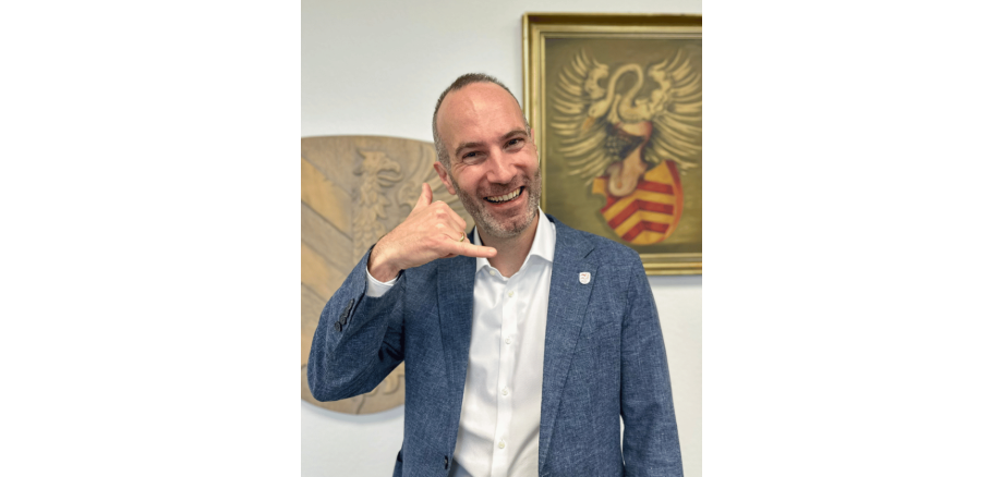 Mann in einem blauen Sakko lächelnd und stehend vor einem Gemälde mit einem Wappen.