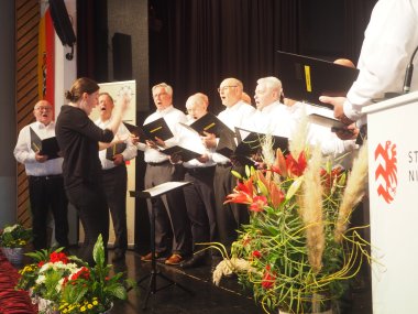 Mehrere Männer stehend in weißen Hemden und schwarzen langen Hosen singend auf einer Bühne. Davor die Dirigentin stehend in schwarz gekleidet.