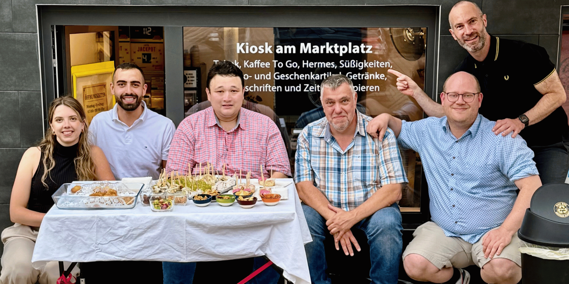 Fünf Männer und eine Frau vor einem Ladengeschäft sitzend. Davor ist ein Tisch mit Fingerfood platziert